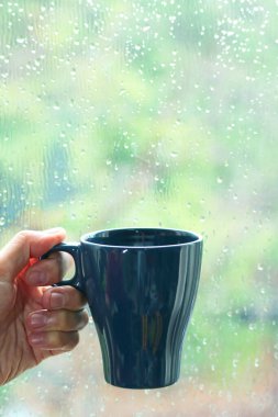 Arka planda yağmurda pencere camı olan bir fincan kahveyi tutarken.