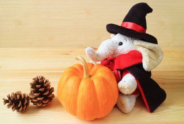 Cute elephant doll in wizard costume with ripe pumpkin and dried pine cones on wooden background clipart