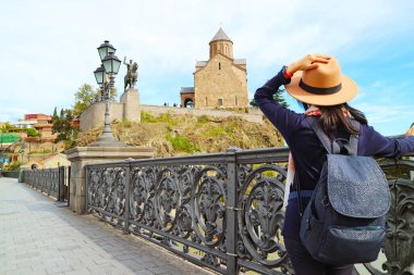 Woman walking on an ornate bridge leading to Metekhi church on the cliff by Mtkvari river, Tbilisi, Georgia clipart