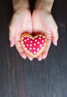 Ellerinde kırmızı ve beyaz bir kalp, koyu siyah ahşap arka planda kraliyet kremalı kurabiye var.