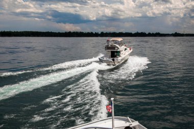  Bir motorlu tekne Kanada 'daki Bin Adalar Gölü' nde köpüklü bir su izi bırakır. Onu mavi gökyüzü altında kabarık bulutlu bir Amerikan bayrağı takip eder.