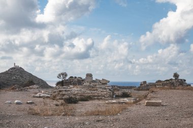 Sahile yakın antik bir arkeolojik alan, sakin bir denizi olan dağınık harabeler ve arkaplanda bir tepede görünen deniz feneri parçalı bulutlu bir gökyüzünün altında..