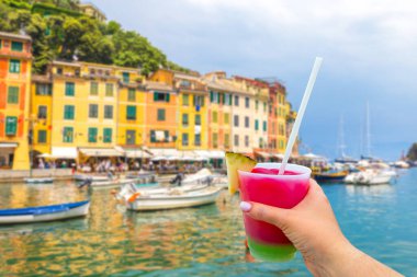 Beautiful Portofino with colorful houses and villas in little bay harbor with Hand holding a glass with cocktail and straw. Liguria, Italy in Europe