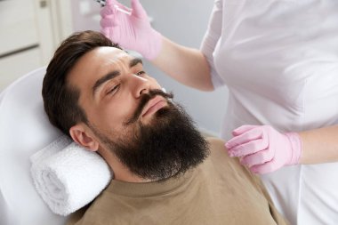 Closeup of bearded man getting beauty injection at aesthetic clinic. Plastic surgeon injecting anti-aging filler in handsome man face. Male cosmetology,