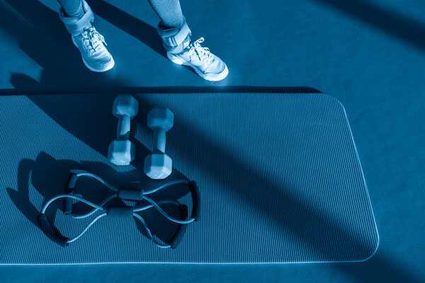 Woman legs near mat with expander and two dumbbells