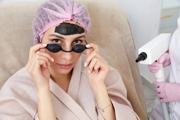 stock image Carbon peeling in beauty salon. Cosmetologist applying black mask on the face of beautiful woman for carbon peeling