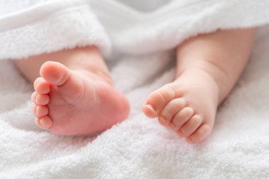 Beyaz bir havlunun lükslü yumuşaklığının altında, bir bebek ayağı ve bacakları belirir, banyo yaptıktan sonra nazik bakımı sembolize eder.