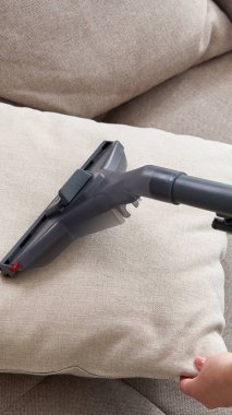 Genç ev hanımı oturma odasında elektrikli süpürgeyle kanepeyi temizliyor.