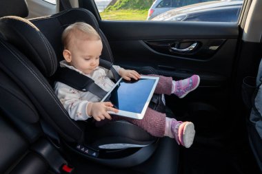 Çocuk oto koltuğunda oturan genç bir kız, uzun bir yolculukta dijital tablet cihazıyla meşgul. Çocuklar için tasarlanmış modern teknoloji ve seyahat konforu kavramı
