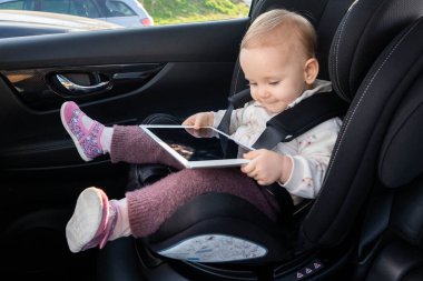 Küçük kız çocuk oto koltuğunda oturuyor, uzun bir yolculuk boyunca eğlenmek için dijital tablet bir cihaz kullanıyor. Çocuklar için eğlenceli ve etkileşimli seyahat deneyimi kavramı