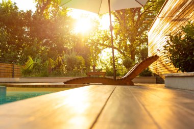 Şık ahşap şezlonglar ve baş döndürücü bir günbatımında yüzme havuzunun yanında beyaz bir şemsiye. Sakin bir açık havada lüks ve konfor kavramı