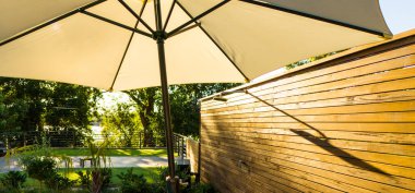 Büyük bir açık hava şemsiyesi ahşap bir çitin yanında gölge, güneş ışığı filtreleniyor ve arka planda yeşillik var. Sakin açık hava rahatlama, mahremiyet ve şık bir gölge kavramı.