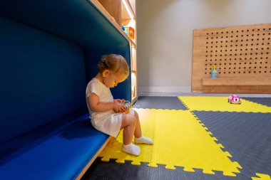 Mavi bir bankta elinde oyuncakla oturan genç bir çocuk. Etrafı renkli ve etkileşimli bir oyun odasıyla çevrili. Erken çocukluk keşfi, eğlence ve yaratıcılık kavramı