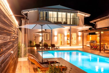 Parlak bir yüzme havuzu, ahşap bir güverte ve geceleri rahat ortam aydınlatması olan lüks bir villa. Rahat ve rahatlamak için modern ve şık bir açık hava yaşam alanı kavramı