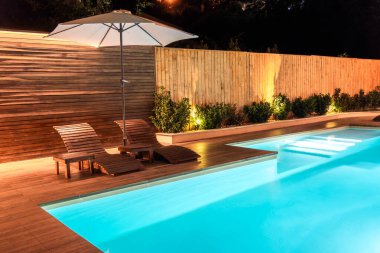 Geceleri rahat ahşap şezlonglar ve villa manzaralı aydınlık bir yüzme havuzu. Lüks açık hava inzivası kavramı