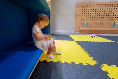 Küçük bir çocuk renkli bir oyun odasındaki rahat bir bankta oturuyor, elinde bir oyuncakla çevreyi keşfediyor. Neşeli oyun anlayışı, erken eğitim ve çocuklar için yaratıcı alanlar