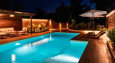 Geceleri ahşap güverteli ve dinlenme odalı lüks yüzme havuzu. Sıcak ışıklandırma ve modern tasarım ile açık alan