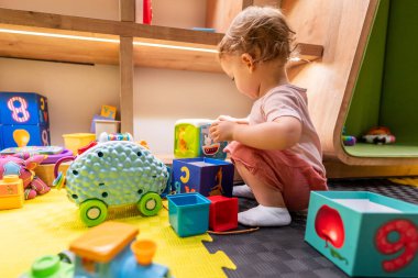 Young child stacking blocks and exploring toys in a colorful indoor playroom clipart