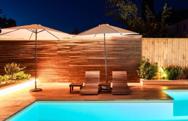 Geceleri şezlonglu sandalyeli ve şemsiyeli şık havuz kenarı. Sıcak ışıklandırmayla aydınlatılmış modern açık hava tasarımı
