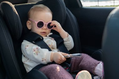 Pembe güneş gözlüklü bir bebek, rahat bir şekilde araba koltuğuna oturmuş, neşeli ve şık bir görünüm sergiliyor. Güvenlik, seyahat ve modaya uygun çocukluk anları.