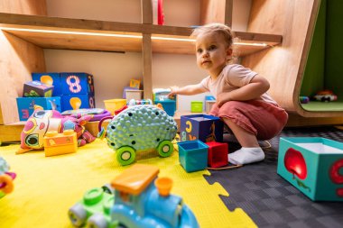 Yeni yürümeye başlayan çocuk hareketli ve neşeli bir oyun alanında eğitim blokları ve oyuncaklarla meşgul.