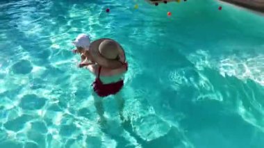 Anne ve bebek güneşli bir günde yeşilliklerle çevrili açık havuzda oyun oynuyorlar..