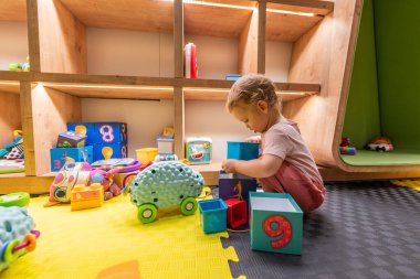 Bebek renkli blokları diziyor ve parlak bir oyun odasında oyuncaklarla oynuyor.