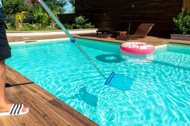 Turkuaz yüzme havuzu temizleyen adam. Kaydırma ağı, ahşap güverte ve renkli şişme şamandıralar.