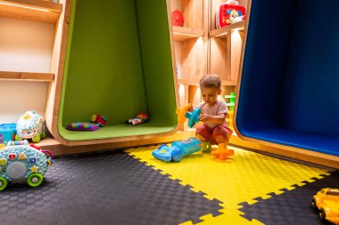 Yeni yürümeye başlayan çocuk öğrenme ve yaratıcılık için tasarlanmış renkli ve modern bir oyun odasında oyuncaklarla oynuyor.