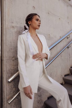 Güzel, şehvetli, siyah saçlı, şık giyimli, zarif beyaz pantolonlu ve ceketli, aksesuarlı bir kadının açık hava fotoğrafı.