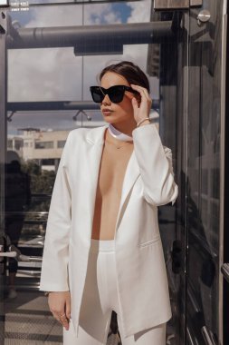 Güzel, şehvetli, siyah saçlı, şık giyimli, şık beyaz pantolonlu ve ceketli, aksesuarlı ve güneş gözlüklü bir kadının açık hava fotoğrafı.