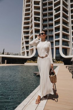 Güzel, siyah saçlı, şık elbiseli ve lüks oteldeki yüzme havuzunun yanında poz veren güzel bir kadının moda fotoğrafı.