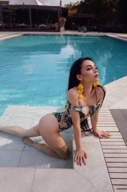 Güzel, siyah saçlı, şık bir mayo giymiş, plaj kulübünde yüzme havuzunun yanında dinlenen güzel bir kadının moda fotoğrafı.