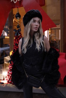 şık kürklü, sarı saçlı ve Noel için dekore edilmiş mağaza vitrinine yakın poz veren güzel bir kadının moda fotoğrafı.