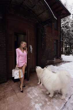 Evin girişine yakın poz veren güzel sarışın bir kadının büyük beyaz bir köpekle oynadığı moda fotoğrafı.