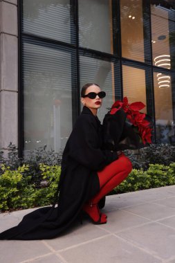 Güzel esmer bir kadının şık elbiseli, siyah ceketli ve aksesuarlı güzel bir buket kırmızı noktalı çiçekle sokakta yürüdüğü bir moda fotoğrafı.