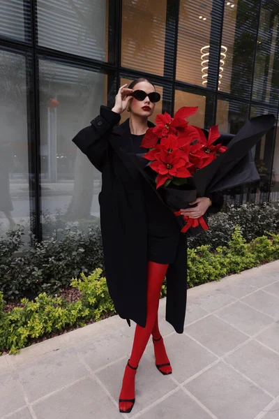 时尚的户外照片 美丽的黑发女人穿着雅致的衣服 黑色外套和饰物 带着一束美丽的红点花在街上走着 — 图库照片