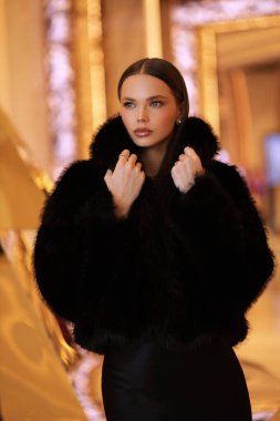 Güzel, şehvetli, siyah saçlı, lüks siyah kürklü ve otel odasında poz veren bir kadının moda fotoğrafı.
