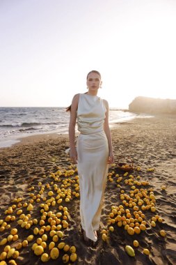 Kıbrıs sahilinde bir sürü limonla poz veren siyah saçlı, zarif beyaz elbiseli güzel bir kadının moda fotoğrafı.