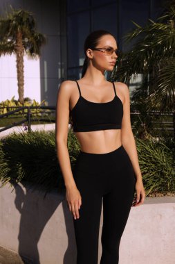 Spor kıyafetli güzel bir kızın yaz bahçesinde poz verirken çekilmiş moda fotoğrafı.