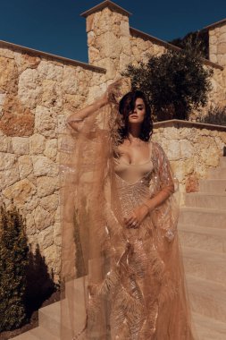 Havalı bej rengi ipek pullu siyah saçlı güzel Slav bir kadının şehir manzaralı güzel bir villada poz verdiği bir moda fotoğrafı.
