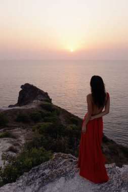 Havalı kırmızı elbiseli ve aksesuarlı, deniz manzaralı tepede poz veren güzel Slav bir kadının açık hava fotoğrafı.
