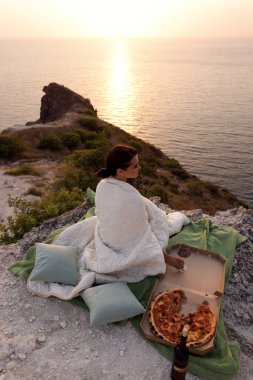 Siyah saçlı güzel bir kadının moda fotoğrafı pizza ve şarapla pikniğe sarılmış gün batımına ve denize bakan bir battaniyeye sarılmış.