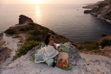 Siyah saçlı güzel bir kadının moda fotoğrafı pizza ve şarapla pikniğe sarılmış gün batımına ve denize bakan bir battaniyeye sarılmış.