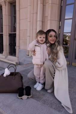 Güzel bir ailenin moda fotoğrafı: uzun sarı saçlı anne rahat beyaz bir palto giyiyor küçük oğluyla sokakta yürüyor
