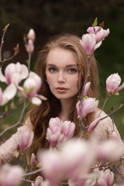 Manolya ağaçlarının yanında poz veren zarif elbiseli, uzun doğal saçlı güzel bir ypung kadının moda fotoğrafı.