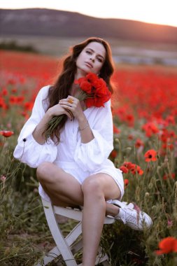 Gün batımında çiçek açan kırmızı gelinciklerle dolu bir tarlada poz veren siyah saçlı güzel Slav bir kadının açık hava fotoğrafı.