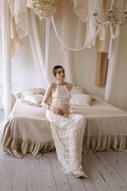 Zarif yatak odasında poz veren siyah saçlı güzel hamile Slav bir kadının moda fotoğrafı.