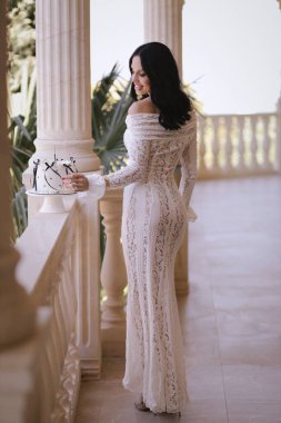 Şık beyaz elbiseli, siyah saçlı güzel bir kadının moda fotoğrafı zarif bir iç mekanda pastayla doğum gününü kutluyor.