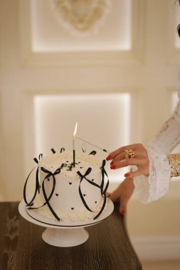 Şık beyaz elbiseli, siyah saçlı güzel bir kadının moda fotoğrafı zarif bir iç mekanda pastayla doğum gününü kutluyor.